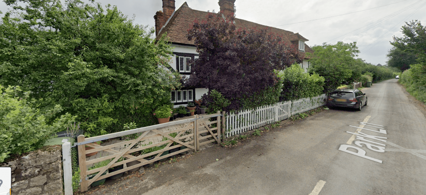 A rural cottage has a white, low-level, painted, timber picket fence, with a five-bar pedestrian gate and larger five-bar gate for vehicle access. Behind the picket fence is an established hedge to provide additional screening and privacy from the road