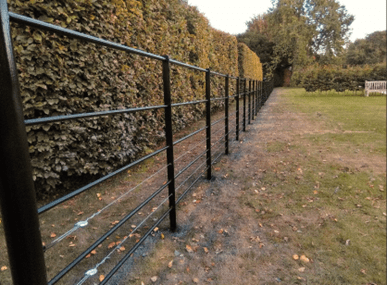 A low-level fence formed of metal bars. The vertical uprights are space between 1.5-2m apart, with 5No. round bars, approximately 30mm in diameter, forming the rails of fence. Within the bottom two rails are additional steel wires 