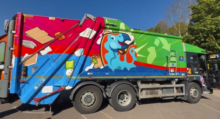 A brightly coloured recycling truck decorated with a cartoon dinosaur and images of recyclable rubbish.