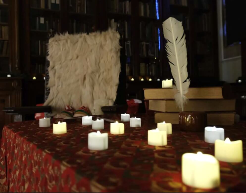 Lit candles on a table next to stacked books, a feather quill, and a fur-trimmed chair in a library setting.